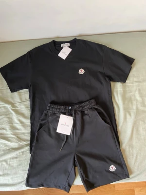 Svarta shorts och T-shirt från Moncler med logga - Säljer ett par stilrena svarta shorts från Moncler med elastisk midja och dragsko. Shortsen har fickor på sidorna och en liten Moncler-logga framtill. Perfekta för sommaren och riktigt sköna att bära till vardags.