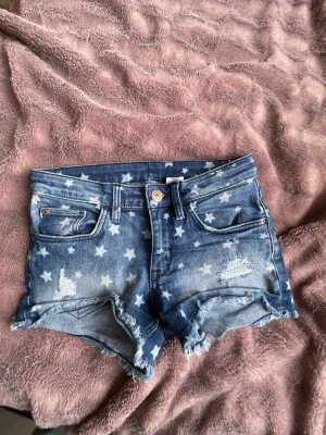 Blå jeansshorts med stjärnmönster - Snygga blå jeansshorts med vita stjärnor och slitna detaljer. Shortsen har rå kant nertill och klassisk femficksdesign. Perfekta för en avslappnad och trendig look.