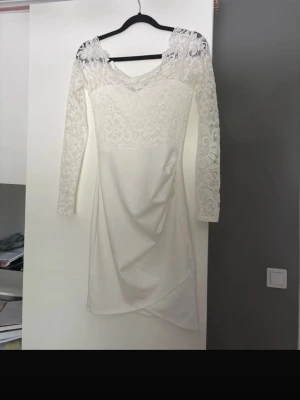 Vit spetsklänning med lång ärm - Säljer en elegant vit kortklänning med spetsdetaljer upptill och på ärmarna. Klänningen har v-ringning, långa spetsärmar och omlott-detalj nedtill. Perfekt för dig som vill ha en romantisk och stilren look.