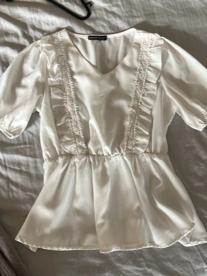 Vit blus med volang  - Söt vit blus i glansigt material med korta puffärmar och volangdetaljer framtill. Blusen har v-ringning och resår i midjan för en feminin siluett. Perfekt att styla med jeans eller kjol för en trendig look.