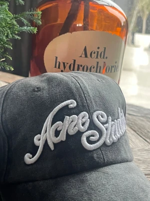 Grå keps från Acne Studios - Säljer en grå keps från Acne Studios med stor vit broderad logga framtill. Kepsen är tillverkad i 100% bomull och har klassisk böjd skärm. Justerbar baktill för perfekt passform. Perfekt accessoar för en trendig streetwear-look.