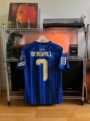 Lucas Bergvall VM 2026 Sverige Bortatröja matchtröja Adidas - Blå matchtröja från Adidas för Sveriges landslag med guldiga detaljer och nummer 7. Namnet 'Bergvall' på ryggen, broderad landslagssköld och FIFA/UEFA-märken på ärmarna. Tröjan har korta ärmar och klassisk v-ringning.