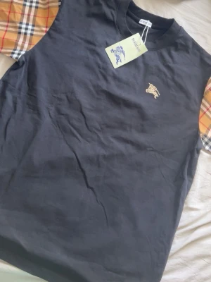 Svart Burberry t-shirt med rutiga ärmar - Snygg svart t-shirt från Burberry med klassiska beige-rutiga ärmar och broderad logga på bröstet. T-shirten har rund hals och är tillverkad i mjuk bomull. Perfekt för dig som vill ha en stilren men ändå unik look. 