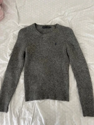 Grå stickad tröja från Ralph Lauren - Mysig grå stickad tröja från Ralph Lauren med rund halsringning och diskret broderad logga på bröstet. Tröjan har långa ärmar och ribbstickade muddar vid ärmslut och nederkant. Perfekt för lager-på-lager och enkel att matcha.