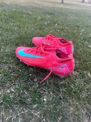 Nike Mercurial rosa fotbollsskor - Säljer ett par Nike Mercurial fotbollsskor i en riktigt snygg rosa färg med turkos swoosh på sidan. Skorna har låg profil, snörning och är tillverkade i ett lätt syntetmaterial som ger bra bollkontroll och grepp på planen. Perfekta för dig som vill sticka ut på fotbollsplanen.