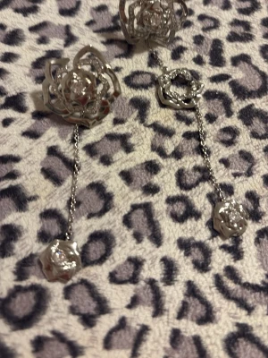 Silvriga rosörhängen från Ioaku - Unika örhängen från Ioaku i form av rosor med hängande kedjor och små rosdetaljer. Örhängena är i silverfärgad metall och har en glittrig finish som ger extra wow-känsla. Perfekta för dig som vill sticka ut med snygga accessoarer.
