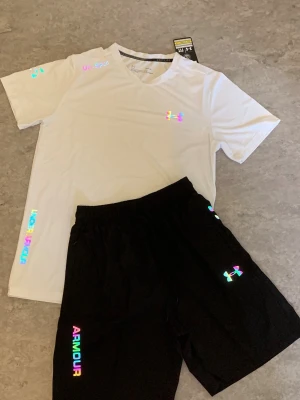 Under Armour träningsset, vit/svart - Snyggt träningsset från Under Armour med en vit t-shirt och svarta shorts. T-shirten har regnbågsfärgade loggor och textdetaljer, rund hals och korta ärmar. Shortsen har elastisk midja och matchande regnbågsfärgad logga och text. Perfekt för gym eller löpning.