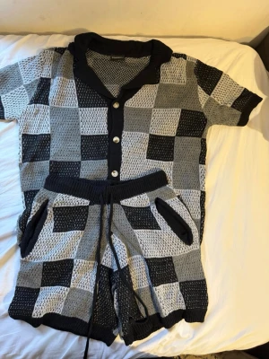 Rutigt set med skjorta och shorts - Sommarn är och varför inte unna sig med perfekt set in för sommaren Snyggt tvådelat set med kortärmad skjorta och shorts i svart, grått och vitt rutmönster. Skjortan har knappar framtill och krage, shortsen har resår och dragsko i midjan samt fickor. Materialet är stickat och luftigt, perfekt för en avslappnad look.