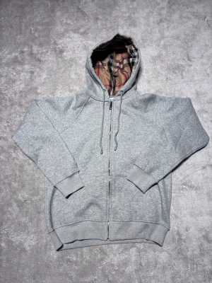 Grå zip hoodie med Burberry-mönstrad huva - Snygg grå zip hoodie från Burberry med klassiskt rutmönstrat foder i huvan. Hoodien har dragsko i huvan, muddar vid ärmslut och nederkant samt två fickor framtill. Perfekt för en chill och stilren look.