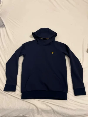 Mörkblå hoodie från Lyle & Scott - Mörkblå hoodie från Lyle & Scott med klassisk gul logga på bröstet. Tröjan har huva, långa ärmar och ribbade muddar vid ärmslut och nederkant. Perfekt för en avslappnad och stilren look.