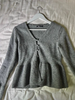 Grå kofta med knappar från Gina Tricot - Mysig grå kofta från Gina Tricot med v-ringning och dekorativa knappar framtill. Koftan har lång ärm och ribbstickad nederdel som ger en snygg siluett. Perfekt att slänga över en topp för en chill look.