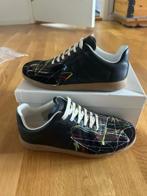 Maison Margiela gats paint splatter 42 - Maison Margiela sneakers i svart läder med färgstänk i rött, gult, blått och vitt. Skorna har vita snören, beige sula och vit insida. Snygg och unik design som sticker ut, perfekt för dig som gillar streetwear och konstnärliga detaljer. Storlek 42, nytt skick