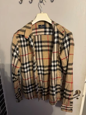 Rutig Burberry skjorta XL - Klassisk rutig skjorta från Burberry i beige, svart, vit och röd. Skjortan har långa ärmar, krage och knappar framtill. Tillverkad i mjuk bomull och har det ikoniska Burberry-mönstret som sticker ut.