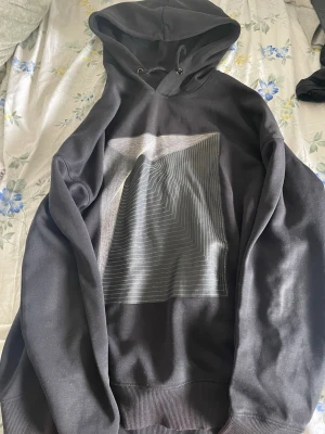 Svart hoodie med grafiskt tryck - Säljer en svart hoodie med huva och dragsko. På bröstet finns ett stort grafiskt tryck i vitt och grått med linjer som bildar ett coolt mönster. Hoodien har ribbade muddar vid ärmslut och nederkant. Perfekt för dig som gillar stilrena och moderna plagg.