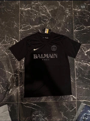 Svart PSG x Balmain t-shirt - Svart t-shirt i sportig modell från Balmain i samarbete med PSG. Tröjan har rund hals, korta ärmar och tryckt Balmain Paris-logga samt PSG-emblem på bröstet. Materialet är troligtvis polyester, perfekt för träning eller streetwear.