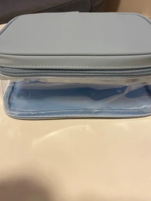 Ljusblå necessär från Apohem - Praktisk och stilren necessär i ljusblått från Apohem Väskan har ett genomskinligt plastmaterial på sidorna och ett mjukt lock med dragkedja. Perfekt för att förvara smink, hudvård eller andra skönhetsprodukter.
