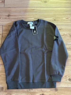 Grå sweatshirt från Stone Island - Snygg grå sweatshirt från Stone Island med klassisk rund hals och ribbade muddar. Tröjan har den ikoniska loggan på vänster ärm och är tillverkad i mjuk bomull. Perfekt för en stilren och avslappnad look.