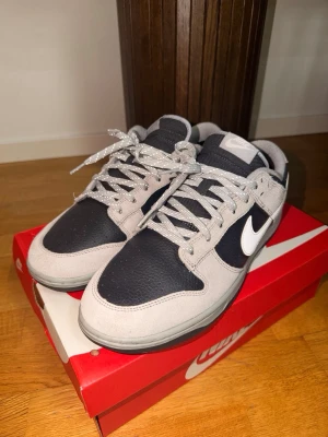 Nike Dunk Low  - Snygga Nike Dunk Low sneakers i grått och svart med vit swoosh. Ovandelen är i mocka och läder, med matchande grå skosnören. Klassisk låg siluett som passar perfekt till streetwear och vardagsstil.