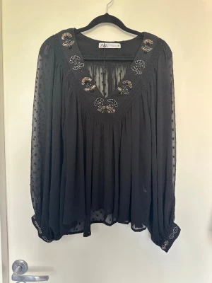 Svart blus med broderi från ZARA Small - Svart, luftig blus från ZARA med transparenta, prickiga långärmar och v-ringning. Dekorerad med fina broderade blommor i beige och guld runt halsringningen och vid ärmsluten. Tillverkad i ett tunt, flowy material som ger en avslappnad look.