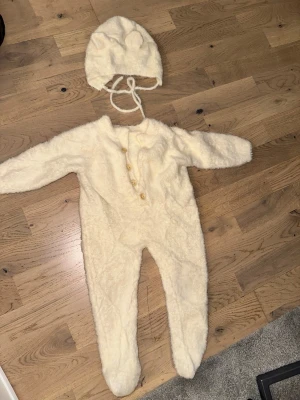 Krämvit fluffig body med mössa - Säljer ett set med en krämvit, supermjuk och fluffig body med matchande mössa. Bodyn har knappar framtill och är helkropp med långa ärmar och ben. Mössan har söta öron och knytband under hakan. Perfekt för att känna sig extra mysig.