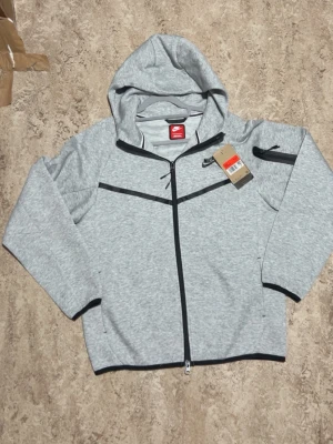 Grå hoodie från Nike med dragkedja - Snygg grå hoodie från Nike med hel dragkedja framtill och svart detalj på bröstet. Hoodien har huva, långa ärmar och en ficka med dragkedja på ena ärmen. Perfekt för en sportig och avslappnad stil.