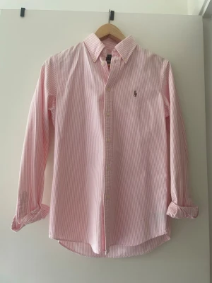 Rosa och vit randig skjorta Polo Ralph Lauren - Klassisk rosa och vit randig skjorta från Polo Ralph Lauren i modellen Custom Fit. Skjortan har lång ärm, knappar framtill och en liten broderad logga på bröstet. Perfekt för en clean och stilren look.