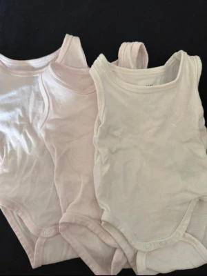3-pack ljusrosa bodys från H&M - Två mjuka bodys från H&M i ljusrosa och offwhite. Bodysarna är ärmlösa med rundad halsringning och tryckknappar i grenen för enkel påklädning. Tillverkade i bomull, perfekta för nyfödda.