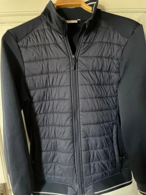 Mörkblå vindjacka med quiltad front - Snygg mörkblå vindjacka med quiltad front och släta ärmar. Jackan har hög krage, dragkedja framtill och två sidofickor med dragkedja. Materialet är en mix av polyester och bomull, vilket ger en sportig och modern look.