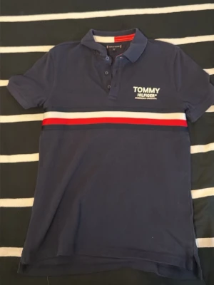 Blå Tommy Hilfiger piké med ränder - Snygg marinblå pikétröja från Tommy Hilfiger med vit och röd rand över bröstet och logga på vänster sida. Klassisk krage och knappar framtill. Perfekt till jeans eller shorts för en clean och sportig look.