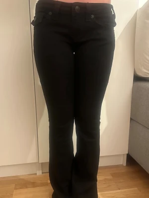 Svarta bootcut jeans från True Religion ”JOEY” - Snygga svarta jeans från True Religion med bootcut passform och väldigt låg midja. I modellen ”JOEY”. Som nya då de bara använts en gång. Säljer pga kommer inte till användning, då jag har andra jeans jag föredrar mer. Jeansen är i storlek W25.