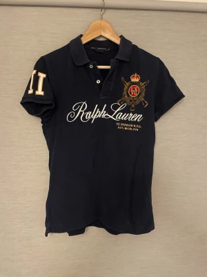 Marinblå pikétröja från Ralph Lauren - Klassisk marinblå pikétröja från Ralph Lauren med broderad logga och emblem på bröstet samt siffran 11 på ärmen. Tröjan har krage, korta ärmar och knappar vid halsen. Perfekt för en sportig och stilren look.
