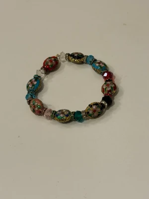 Flerfärgat armband med blommor - Handgjort armband med ovala pärlor i olika färger som turkos, röd, svart och rosa. Varje pärla har detaljerade blommönster och guldfärgade kanter. Armbandet är elastiskt och har en bohemisk vibe 