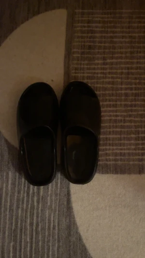 Svarta slip-in tofflor i plast - Säljer ett par svarta slip-in tofflor i plast med enkel och clean design. Tofflorna har rundad tå och öppen häl, perfekta att glida i hemma eller på gymmet. Inga synliga märken eller loggor. Lätta att hålla rena och supersmidiga att använda.