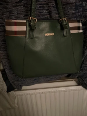 Grön handväska från Burberry Äkta - Stilren grön handväska från Burberry med klassiskt rutmönster på sidorna och guldfärgade detaljer. Väskan har två rejäla handtag och är tillverkad i ett slätt konstlädermaterial. Perfekt för dig som vill ha en tidlös och trendig accessoar. Den är lite sönder vid sidan. Normalt så kostar den här 8000 men jag säljer min för 2500 pris kan diskuteras 