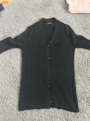 Svart ribbad kofta med knappar - Snygg svart ribbad kofta från Gina Tricot med v-ringning och långa ärmar. Koftan har en smal passform och stängs med fem svarta knappar framtill. Perfekt att slänga över en topp för en stilren look.