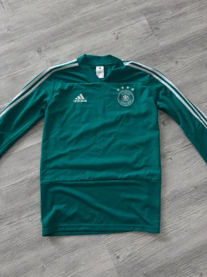Grön långärmad Adidas fotbollströja - Snygg grön långärmad fotbollströja från Adidas med vita detaljer och tre ränder på ärmarna. Tysklands landslagsemblem med fyra stjärnor på bröstet. Perfekt för träning eller match. Tillverkad i lätt och funktionellt material. Storlek S