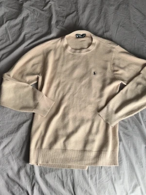 Beige stickad tröja från Polo Ralph Lauren - Klassisk beige kashmir tröja från Polo Ralph Lauren med rund halsringning och diskret broderad logga på bröstet. Tröjan har långa ärmar och ribbade muddar vid ärmslut och nederkant. Perfekt för lager på lager och enkel att matcha. Skriv om ni har några fler frågor om tröjan, priset är ej hugget i sten