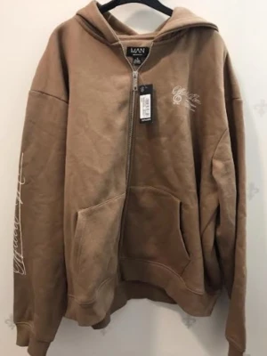 Brunt set med hoodie och byxor - Säljer ett brunt mjukisset från BoohooMAN med dragkedja på hoodien och fickor framtill. Setet har vita tryckdetaljer på bröstet och ärmen samt på byxan. Byxorna har resår i midjan och är raka i modellen. Perfekt för en avslappnad och trendig look.