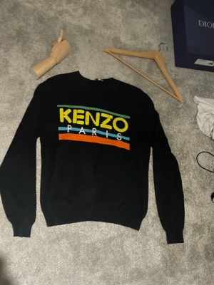 Svart stickad Kenzo-tröja unisex - Säljer en svart stickad tröja från Kenzo Paris med färgglatt tryck i gult, grönt, blått och orange på bröstet. Tröjan har rund halsringning och ribbade muddar vid ärmslut och nederkant. Perfekt för dig som gillar streetwear och vill sticka ut. Passar hundra procent M och S! Kom med ett pris!