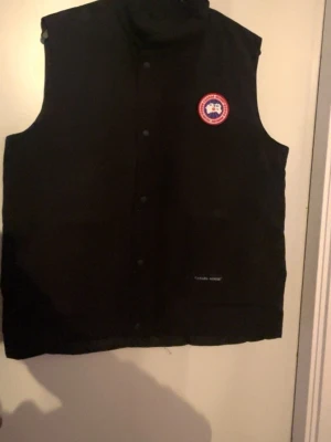 Svart dunväst från Canada Goose - Snygg svart dunväst från Canada Goose med klassisk logga på bröstet och diskret varumärkesbrodyr vid fickan. Västen har hög krage och knäppning med tryckknappar. Perfekt för lager-på-lager och håller dig varm under kyliga dagar.