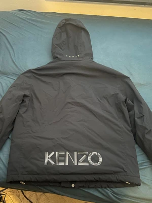 Svart vindjacka från Kenzo - Snygg svart vindjacka från Kenzo med stor logga på ryggen med reflex och insidan. Jätte eftersökt model. Jackan har huva och dragsko nedtill för extra passform. Materialet är tunt och vindtätt, perfekt för blåsiga dagar. Stilsäker design med tydlig streetwear-känsla.