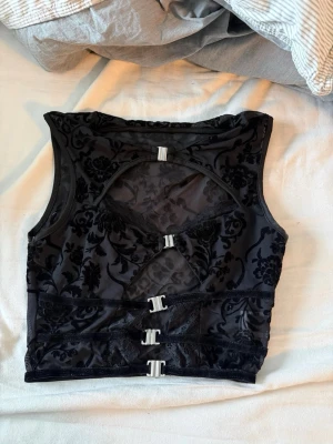 Svart mesh topp med sammetsmönster - Snygg svart topp i mesh med sammetsmönster och dekorativa spännen framtill. Kroppen har ett genomskinligt tyg med blommigt mönster och flera silvriga spännen som ger en edgy look. Perfekt för dig som gillar detaljer och vill sticka ut.