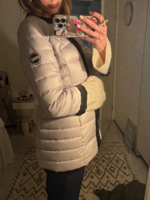 Ljusbeige pufferjacka från Colmar - Snygg ljusbeige pufferjacka från Colmar med svarta detaljer längs dragkedjan och ärmsluten. Jackan har en quiltad design och lång ärm, perfekt för kyliga dagar. Märkeslogga på ärmen ger en sportig touch. Passar till dig som vill ha en stilren och varm jacka.