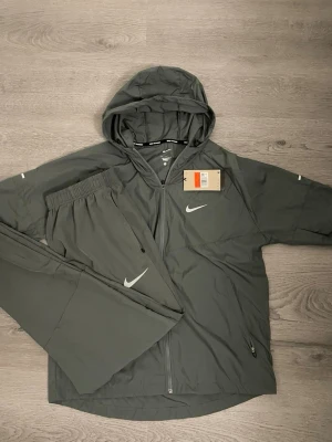 Grå träningsset från Nike - Snyggt grått träningsset från Nike med jacka och byxor. Jackan har huva, dragkedja framtill och två fickor. Byxorna har resår i midjan och diskret Nike-logga. Perfekt för träning eller chill. Materialet är lätt och känns skönt mot huden.