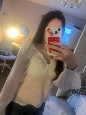 Prickig vit blus med volang från Zara - Söt vit blus med diskret prickigt mönster, volang vid axlarna och spetsdetaljer längs urringningen. Långa ärmar med breda muddar och lätt transparent material. Perfekt för en trendig och romantisk look.