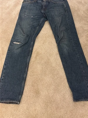 Tiger of Sweden - Säljer ett par blå jeans med rak passform och storlek W29 L32 och ett snyggt hål på vänster knä. Jeansen har klassisk design och är gjorda i denim. Perfekta för en avslappnad och trendig look. Om ni har frågor så tveka inte på att höra av er