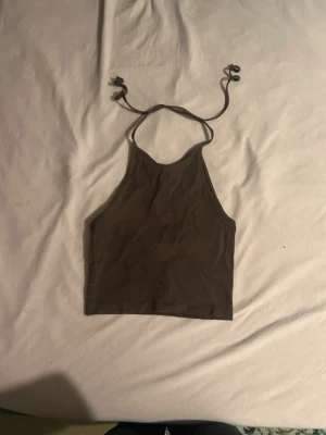 Brun croppad linnetopp med knyt - Säljer en brun croppad linnetopp med tunna axelband som knyts i nacken. Toppen har en enkel och stilren design med hög halsringning och ärmlös passform. 