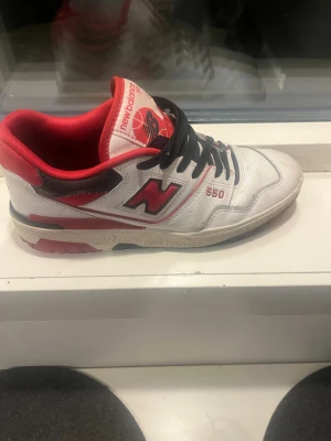 New Balance 550 vita/röda sneakers - Säljer ett par New Balance 550 sneakers i vitt med röda och svarta detaljer. Skorna har svart snörning, röd insida och klassisk NB-logga på sidan. Ovandelen är i läder och sulan har röda och svarta inslag. Perfekta för dig som gillar retrostil.