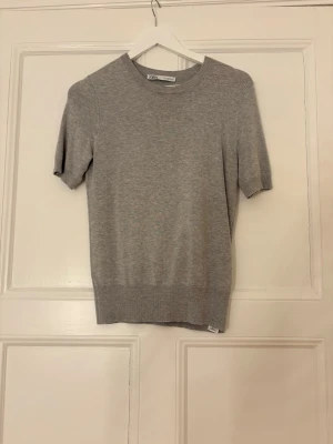 Grå stickad t-shirt  - Snygg grå stickad t-shirt med rund halsringning och korta ärmar. Tröjan har en minimalistisk design och är tillverkad i mjukt, tunt stickat material som passar perfekt till lager på lager eller för en clean look. Den har blivit lite nopprig. Den är i M men sitter som en S