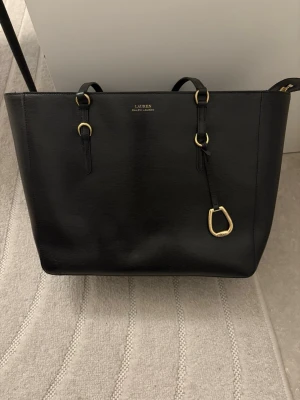 Svart shoppingväska från Lauren Ralph Lauren - Stilren svart shoppingväska i slätt läder från Lauren Ralph Lauren. Väskan har två axelband, guldfärgade metalldetaljer och en dekorativ hängare framtill. Perfekt för dig som vill ha plats för allt och gillar klassisk design.
